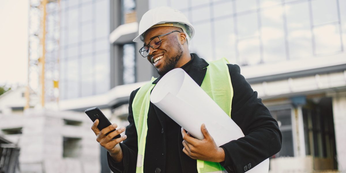 Young black race man with blueprint stading near glass building. Talking by phone.